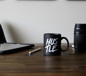 desk with laptop, camera, and mug printed with "hustle"