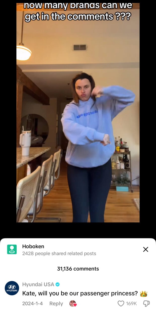 A woman in a blue hoodie performs a dance in a kitchen with text saying “how many brands can we get in the comments ???” Hyundai USA comments, “Kate, will you be our passenger princess?” with a crown emoji.