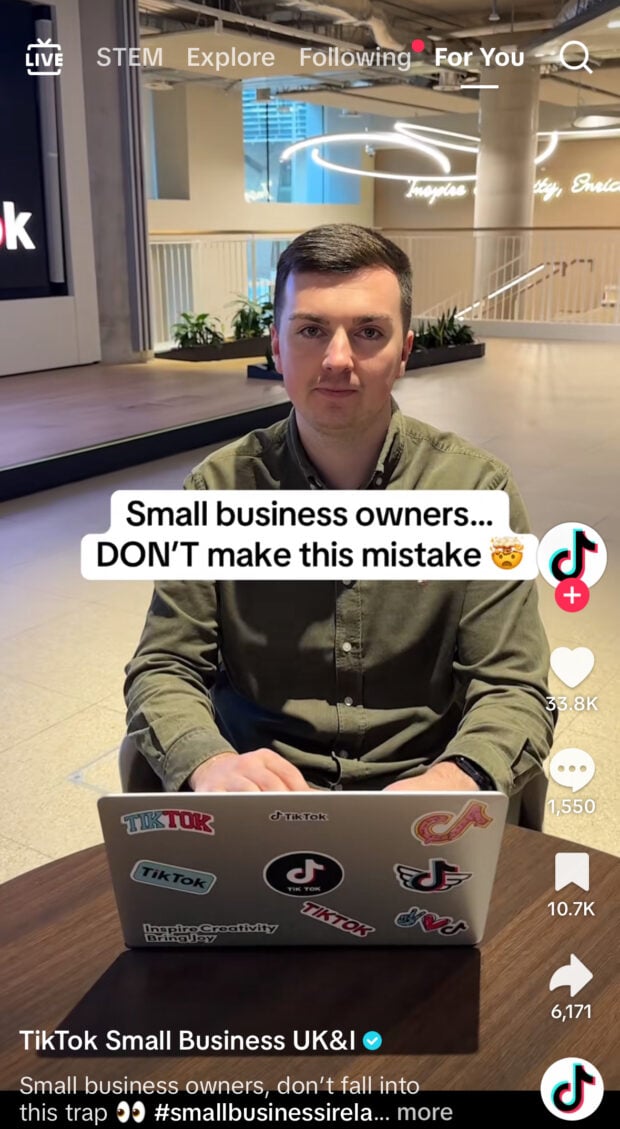A man sits at a table with a laptop covered in TikTok-themed stickers. The overlay reads “Small business owners… DON’T make this mistake 🤯” inside a TikTok HQ office.