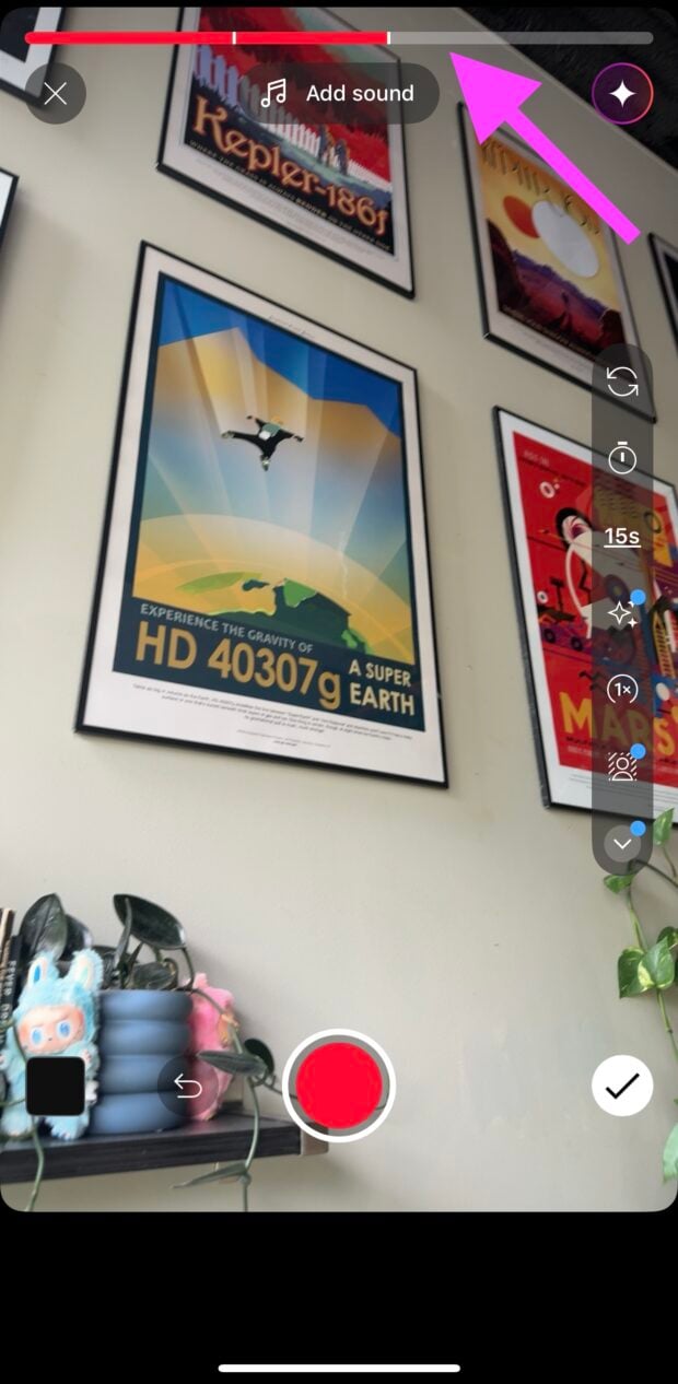 YouTube Shorts recording screen with a pink arrow pointing to the top progress bar. Framed space-themed posters are on the wall, with a shelf of books, plants, and plush toys visible at the bottom