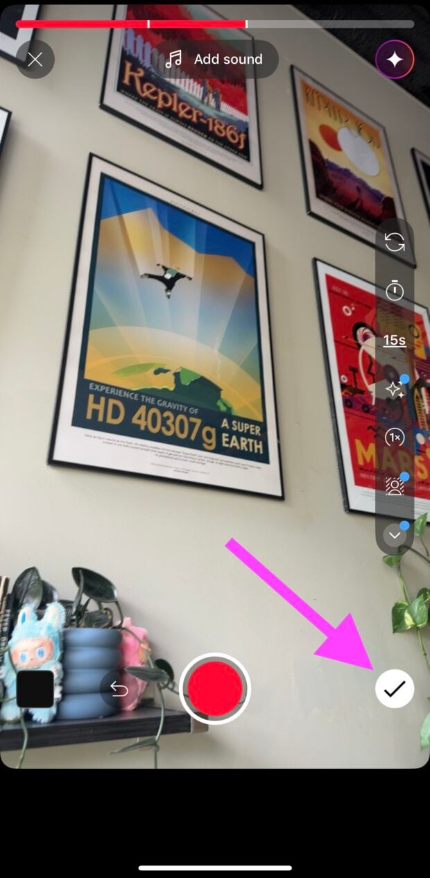 YouTube Shorts recording interface with a pink arrow pointing to the white checkmark button in the lower-right corner, indicating the next step. A progress bar is visible at the top, with space-themed posters on the wall and plush toys on a shelf below