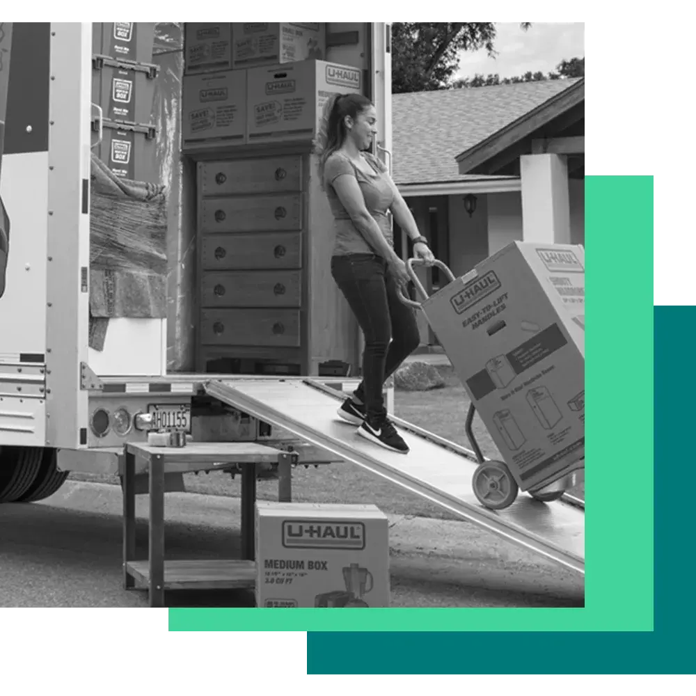 A black and white photo of a woman on a ramp, pushing a hand truck with a U-Haul box on it into the back of a moving truck.