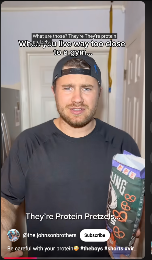 Screenshot of a YouTube Short by @the.johnsonbrothers. A man in a black t-shirt and backwards cap holds a bag of protein pretzels. On-screen text reads: "When you live way too close to a gym… They're Protein Pretzels." Caption: "Be careful with your protein #theboys #shorts."
