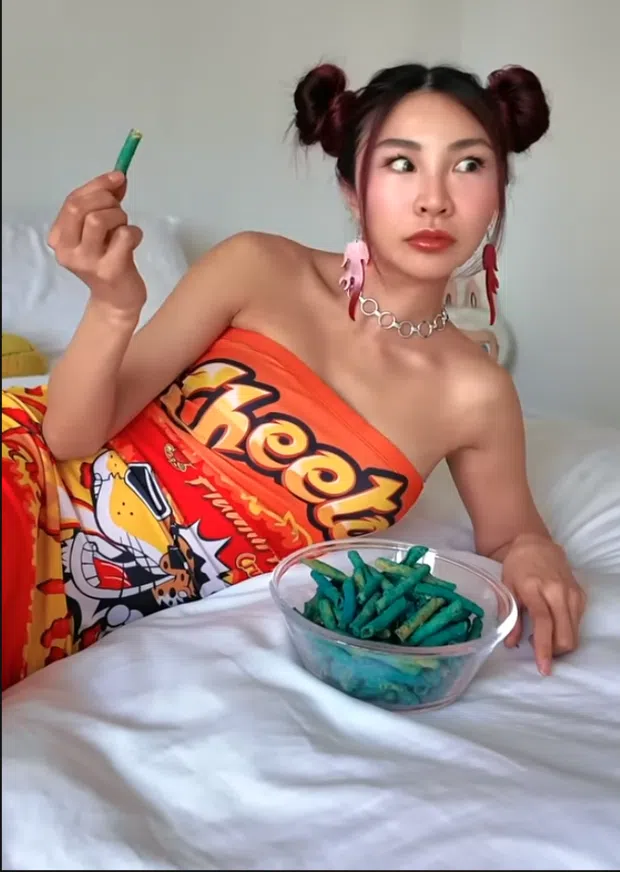 Screenshot of a video by an unidentified creator. A woman in a Cheetos-branded tube top reclines on a bed, holding a green snack and looking at a bowl of green puffed snacks. No brand or platform handle visible.